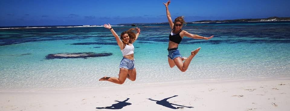 Two girls jumping one the beach. Beach has white sand and blue water.