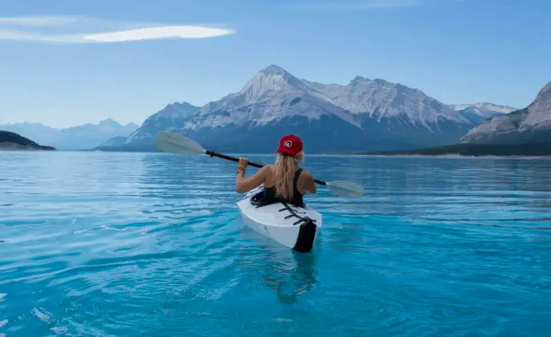 A girl is in a canoe on the water. Blue water in the foreground and mountains in the background.