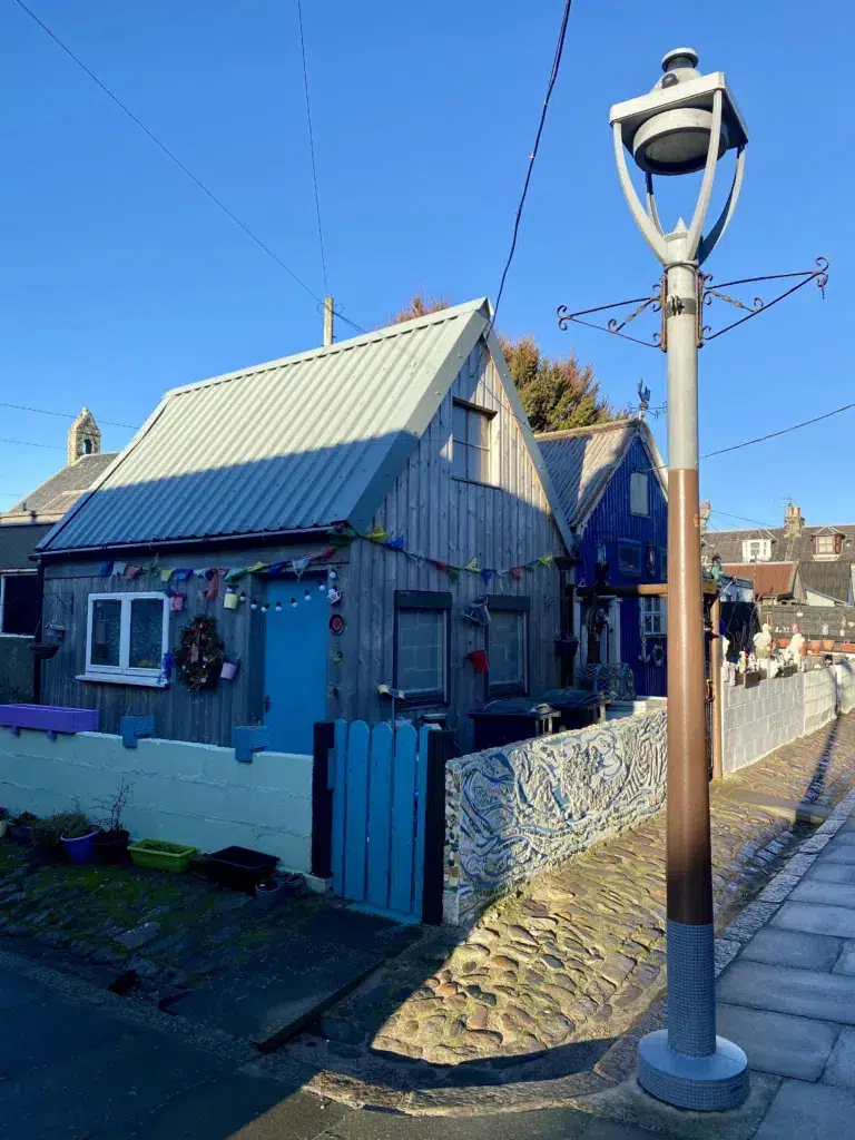 An old fashioned lamp post in the right foreground. A blue shed and gare in the background in Footdee.